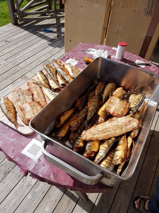 Stor metallbytta fylld med varmrökt fisk och kött på ett träbord utomhus, med kryddburk och verktyg i bakgrunden.