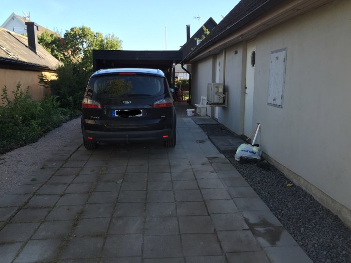 Bil parkerad på uppfart bredvid hus och carport, grå plattor och grus bredvid uppfarten.