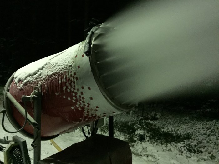 Snökanon i drift som skapar snö på natten med omgivande snötäckt mark.