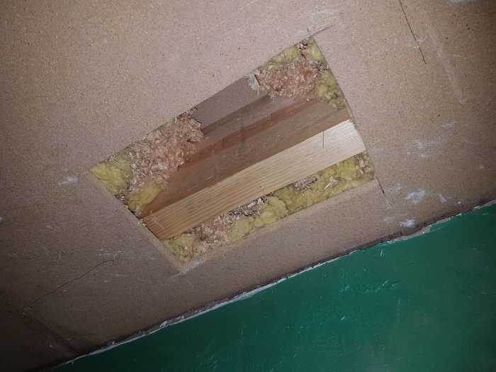 Hål i innertak som visar gips, spånskiva, mineralull och en träbjälke, indikerar vägg konstruerad efter taket.