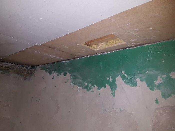 Hål i innertaket som visar lager av gips, spånskiva och synlig bjälke med bortskrapad mineralull och en vägg som fortsätter under taket.