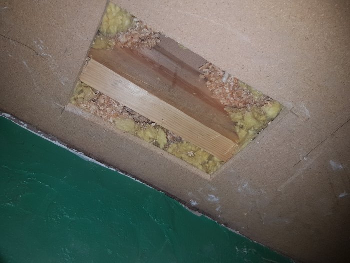 Öppning i ett innertak som visar en gips- och spånskiva, mineralull och en bjälke, med tecken på annat rums konstruktion.