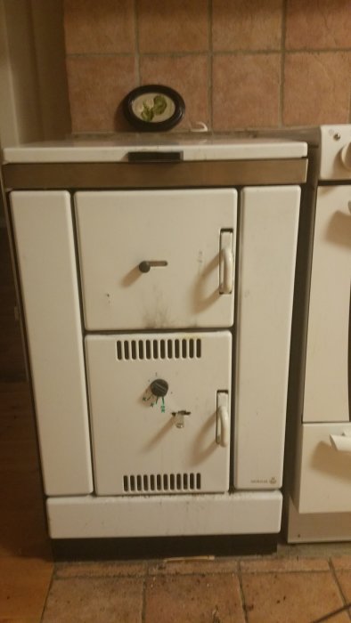 En vit Wamsler vedspis i ett kök med tydliga bruksspår och en klocka ovanpå.