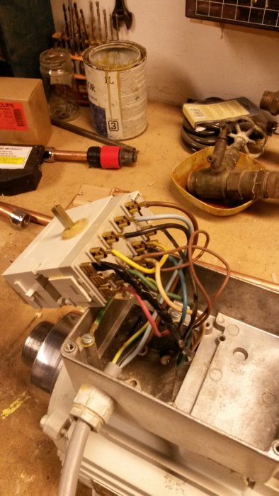 Öppnad vit elektrisk kopplingsbox med exponerade kablar och terminaler på ett verkstadsbord med verktyg i bakgrunden.