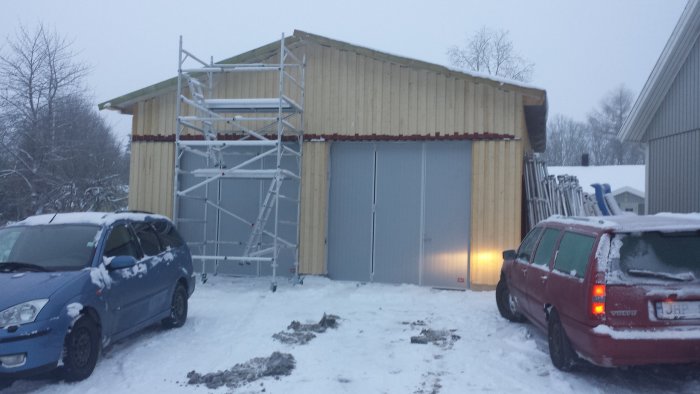 Nybyggd träpanelklädd byggnad med ställning och två bilar i snöig omgivning under skymning.