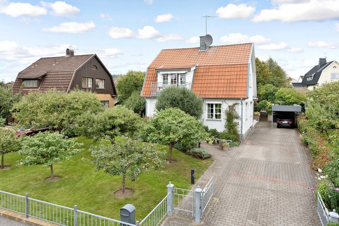 Framsida av vitt hus med tegeltak, omgivet av grönskande trädgård och infart med bil i carport.