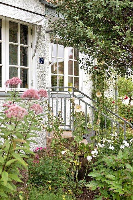 Vardaglig framsida av hus med trädgård och entrétrappa omgiven av blomster och grönska.