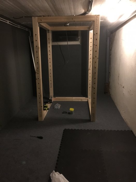 Hemmagjort powerrack av träreglar i en källarutrymme med verktyg och skyddsmatta på golvet.