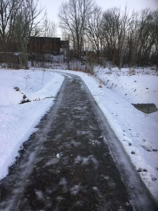 Smal, isig väg mellan snötäckta diken, mätt från kant till kant, omgiven av bar trädvegetation.