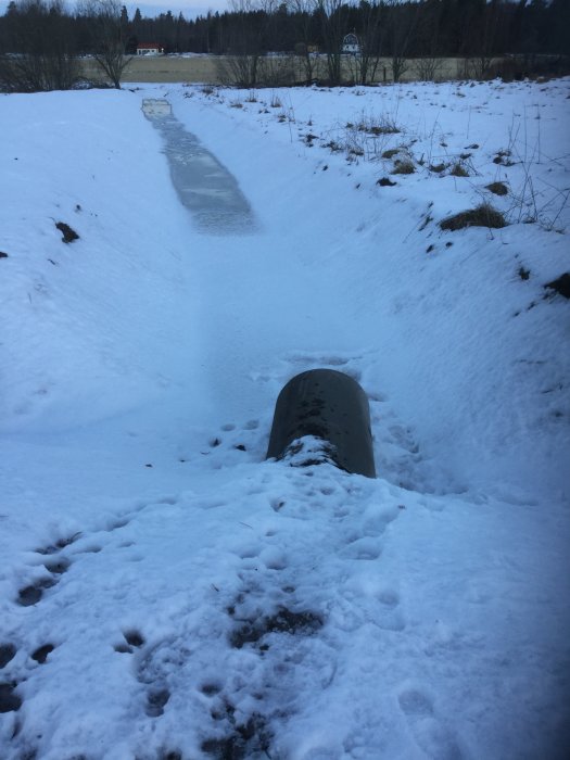 Delvis snötäckt avloppsrör i landskap med synlig isbildning och angränsande snötäckt mark.