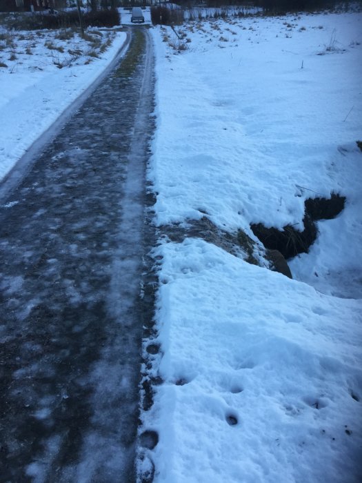 Snötäckt grusväg med hjulspår och en bil i bakgrunden, gränsande till ett tomt snöfält.