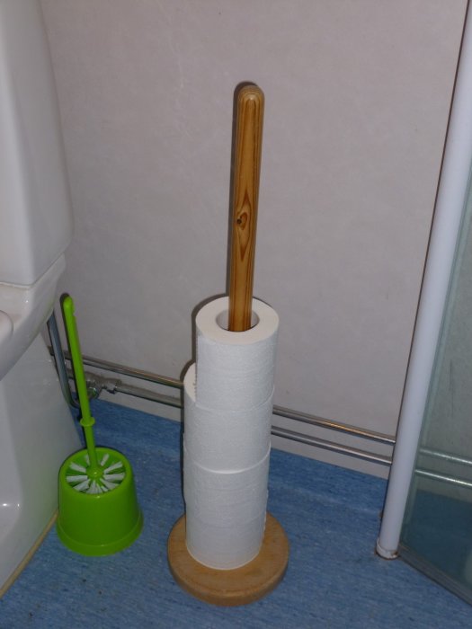 Toarulle på vertikalt hållare med tre lager, bredvid grön toalettborste, i ett badrum.