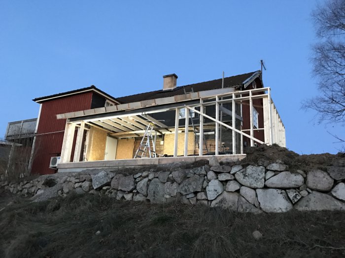 Hus med pågående renovering och nybyggd veranda i trä mot en blå himmel vid skymningen.