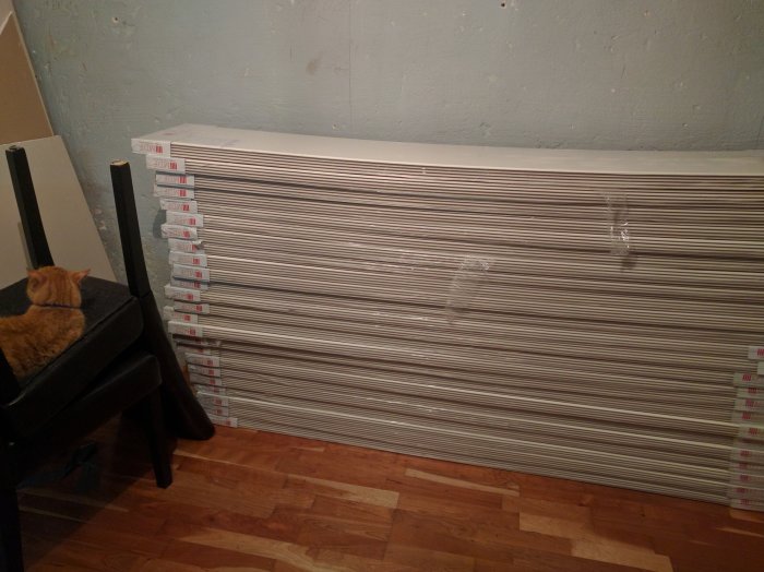 En stapel med nya panelbrädor inplastade bredvid en stol med en röd katt sittande på det mörka tyget.