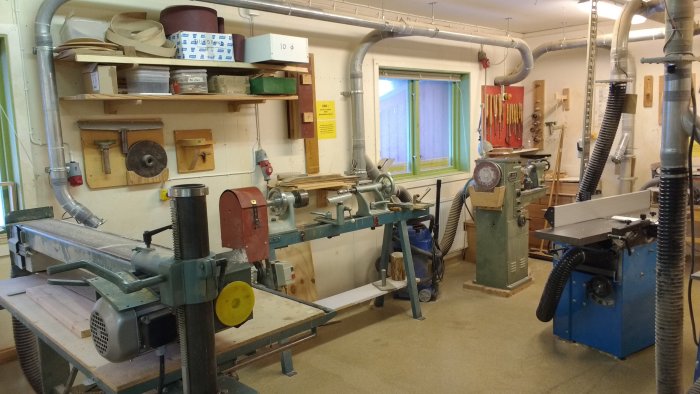 Välutrustat snickeriverkstadsrum med en svarv, bandslip, fräsbord och planhyvel omgivna av hyllor med verktyg och material.