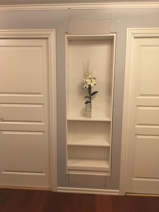 Vit inbyggd hylla i vägg med tre hyllplan, det översta med en vas och vita blommor, omgivet av klassisk panel.