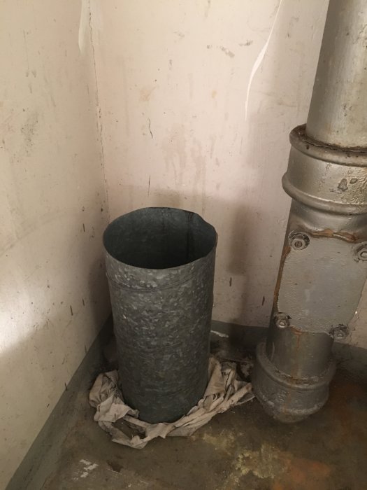 Ett metallrör som går upp från golvet bredvid ett större rör i ett gammalt pannrum, omringat av trasor vid basen.