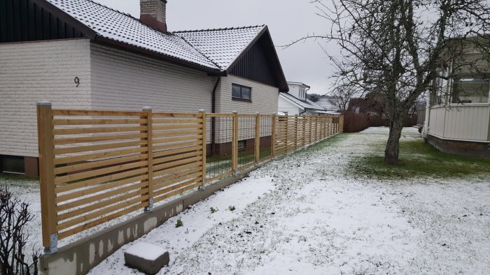 Nybyggt trästaket med stolpar fästa i stolpskor och expanderbultar, på snöbeklädd tomt.