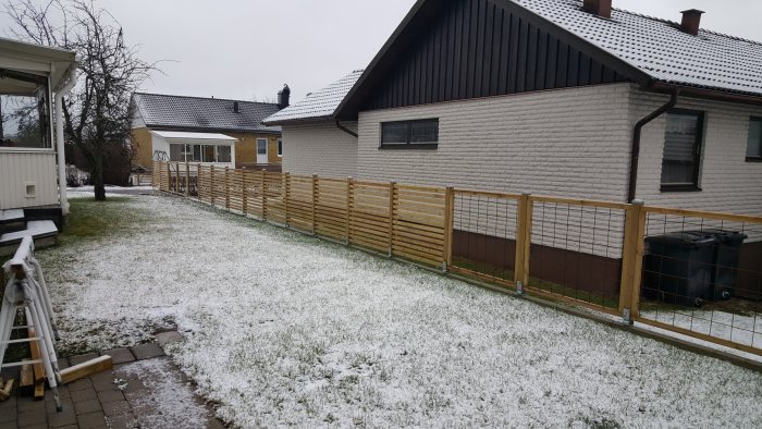 Nyinstallerat staket med träribbor och metallnät på en snötäckt gräsmatta intill ett tegelhus.