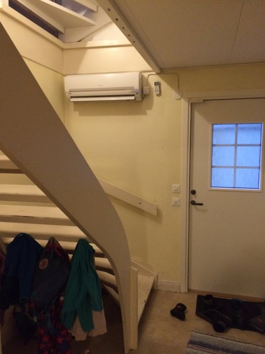 Innerrum med en vit trappa och förborrade hål för spaljé, en dörr och en AC-enhet på väggen.
