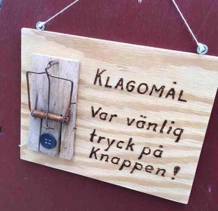 Träskylt med texten "KLAGOMÅL var vänlig tryck på knappen!" och en råttfälla monterad som knapp.