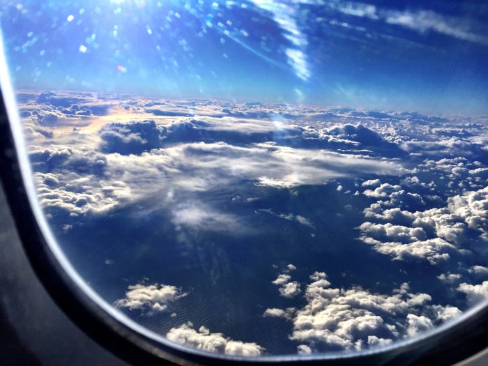Flygplansfönstervy av blå himmel, lysande sol, fluffiga moln och ljust havsskimmer.