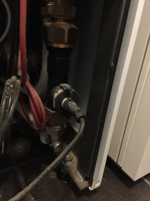Svart komponent med läckage vid anslutningar i ett värmesystem, omgiven av kablar och rör.