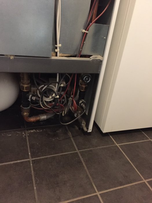 Läckage vid svart komponent under en värmepanna, med synliga rör och ledningar.