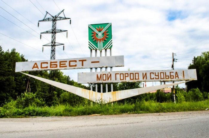 Vägskylt med texten "Асбест - мой город и судьба!" och staden Asbests emblem framför kraftledningsstolpar.