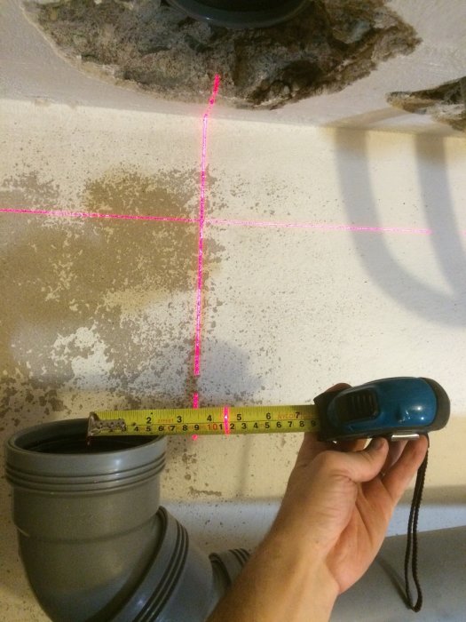 Hand holding a tape measure against PVC pipes with a laser level projecting a cross on a mottled wall during installation.