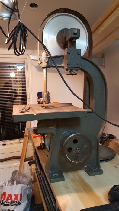 Gammal bandsåg i gjutjärn på träbänk, med synliga remskivor och bandsågshjul, före renovering.