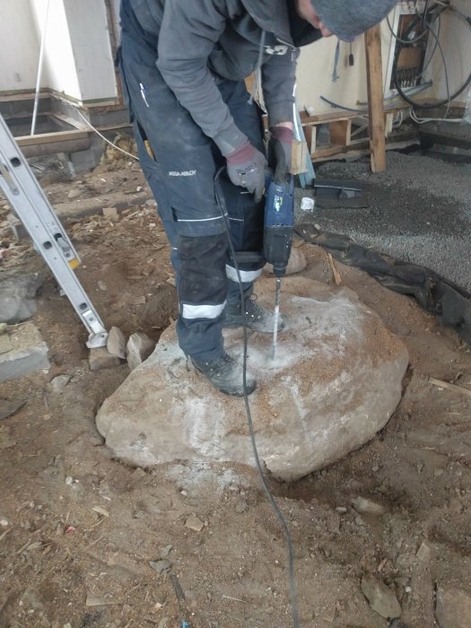 Person borrerar hål i en stor sten på krypgrund med en handhållen borrmaskin.