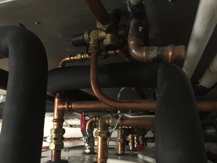 En komplex installation av rör och ventiler under en varmvattenberedare, med synligt skvallerrör.