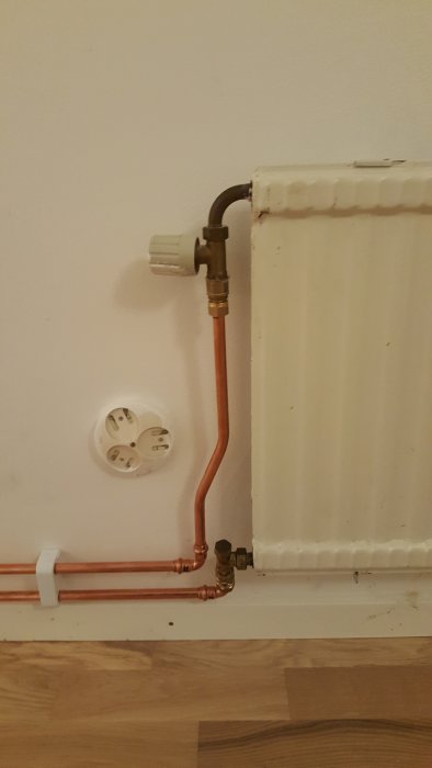 Installationsvy av en radiator med kopparledningar och en närliggande strömuttag på en vägg.