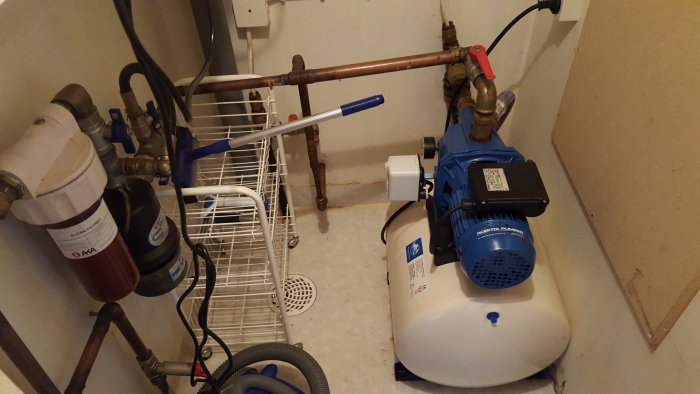 Pumpautomat med blå motor och vit vattenbehållare i ett pumprum med kopparledningar.
