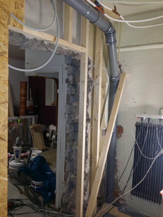 Orenoverat rum med exponerade väggstommar, synliga rörinstallationer och elledningar mot en bakgrund av en delvis riven vägg.