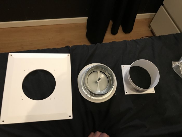 Delar till en frånluftsventil utspridda på ett mörkt tyg med golv och gardiner i bakgrunden.