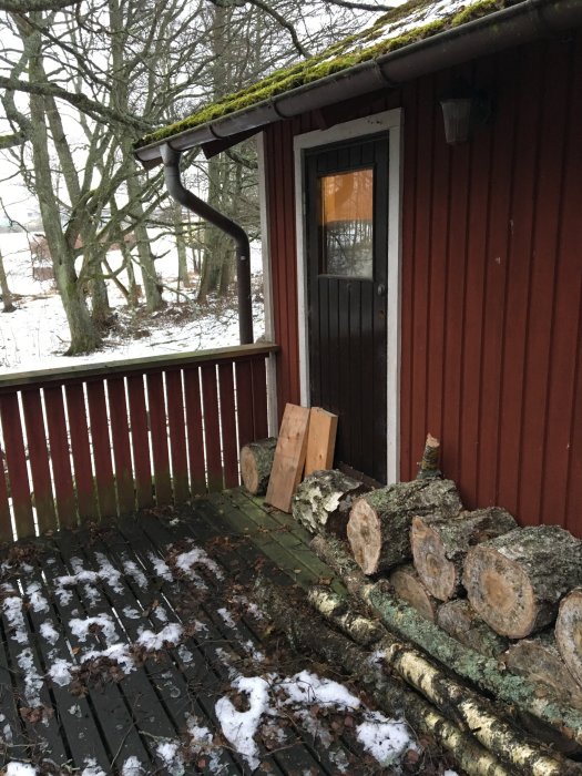 Rödfärgad snickarbod med öppen dörr, omgivet av trästockar och lätt snötäcke på trädäcket.