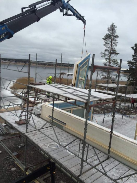 Byggnadsarbetare monterar en väggsektion med en kran på byggarbetsplats vid vattnet.