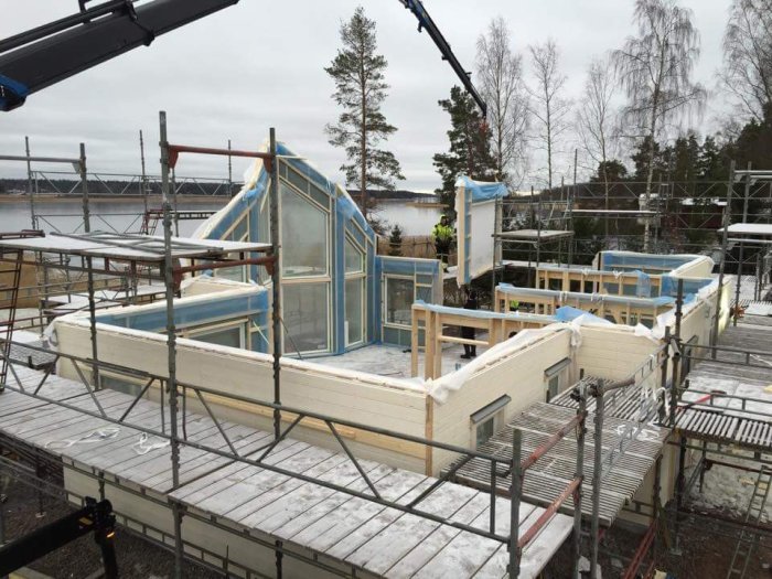Montage av ett hus med synliga vitmålade väggar och blå skyddsfilm på fönsterna, vid sjökant med skog i bakgrunden.