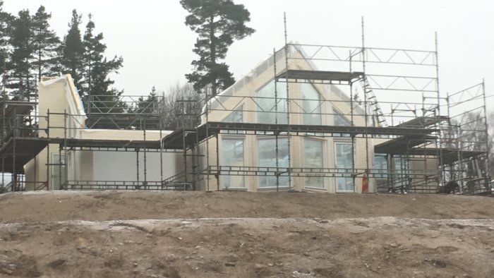 Ett hus under konstruktion, omgivet av byggnadsställningar, med synliga skivmaterial och fönsterpartier.
