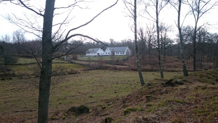 Vy över en lantlig fastighet med ett vitt hus omgivet av öppna gräsmarker och lövträd, med stenmurar som markerar avgränsningen.