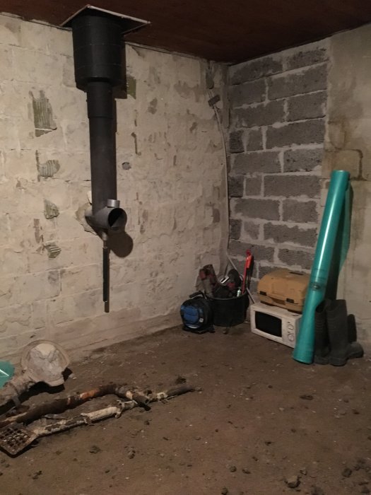 Svart kaminrör som sticker ut från en källarvägg med verktyg och byggmaterial på golvet.