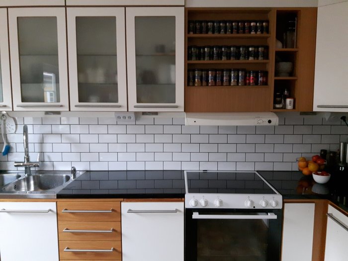 Kök med vita skåp, svart bänkskiva, vit kakelvägg och öppen hylla med burkar.