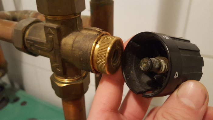 Hand håller en demonterad radiatorventil med fjädermekanism, bredvid mässingskoppling på rör.