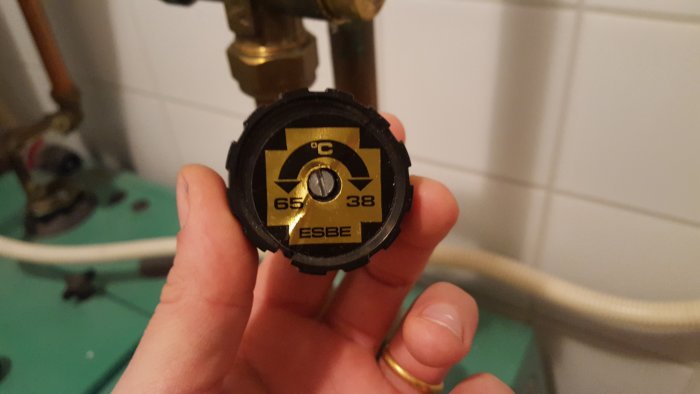 Hand håller en demonterad ESBE-termometerinställning med tydliga temperaturmarkeringar mot en suddig bakgrund av rör och badrumskakel.