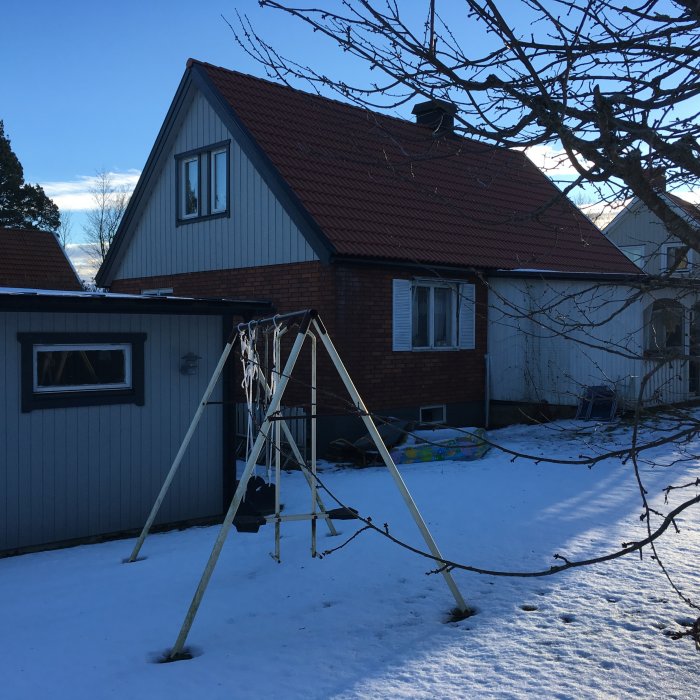 En villa med sadeltak och tegelvägg, en del klädd i träpanel, med snöig trädgård och gungställning i förgrunden.