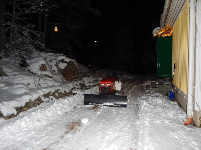 En röd snöslunga med frontmonterat snöblad på en snötäckt väg vid sidan av ett hus och stenmur i skymningen.