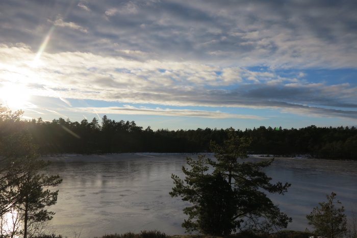 Solnedgång över frusen sjö omgiven av skog och moln på himlen.