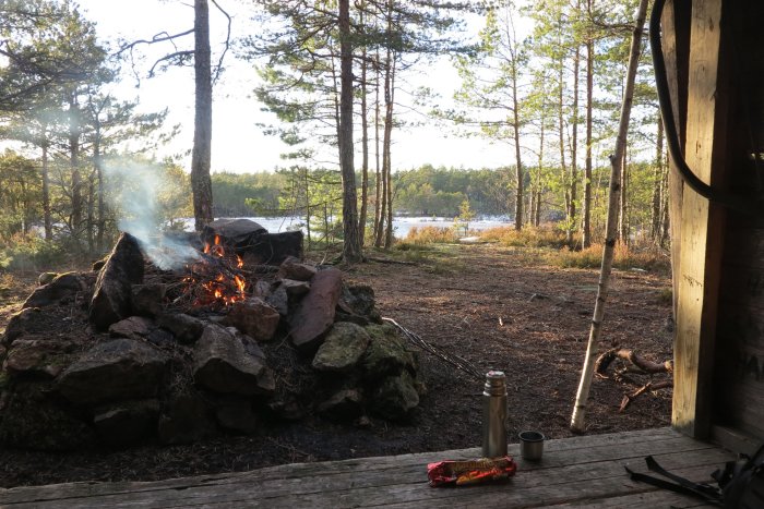 Eldstad utomhus med brinnande eld vid sjö, termos och kexchoklad på träbänk.
